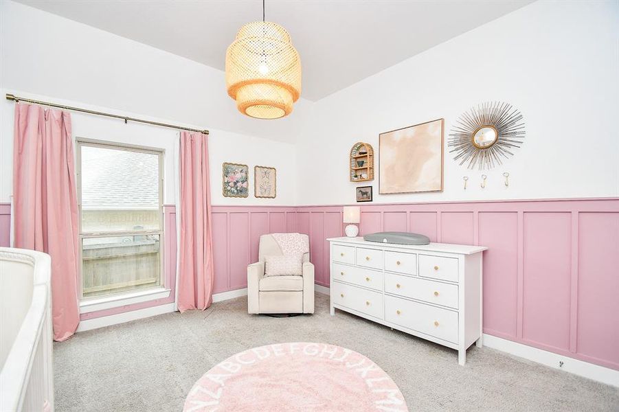 This is a cozy, well-lit nursery room with a white and pastel pink color scheme, featuring wainscoting. It includes a window dressed in pink curtains, providing natural light.