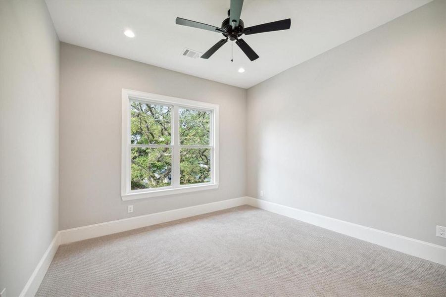 This is a bright, empty room featuring neutral walls, carpeted flooring, a large window with a view of trees, and a ceiling fan with lighting.
