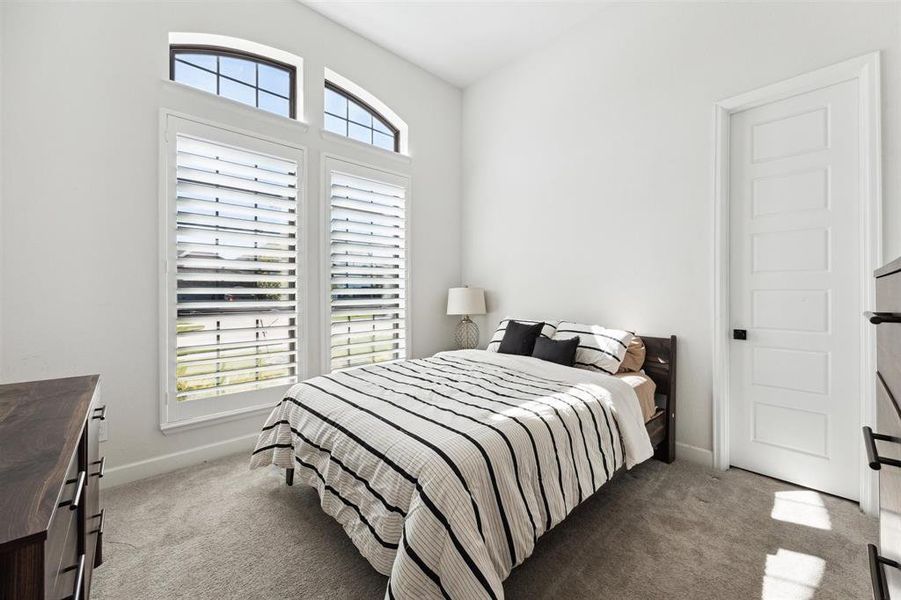 A bright and airy  secondary bedroom features large arched windows with plantation shutters, a plush carpet, and neutral color tones, creating a serene and spacious atmosphere.