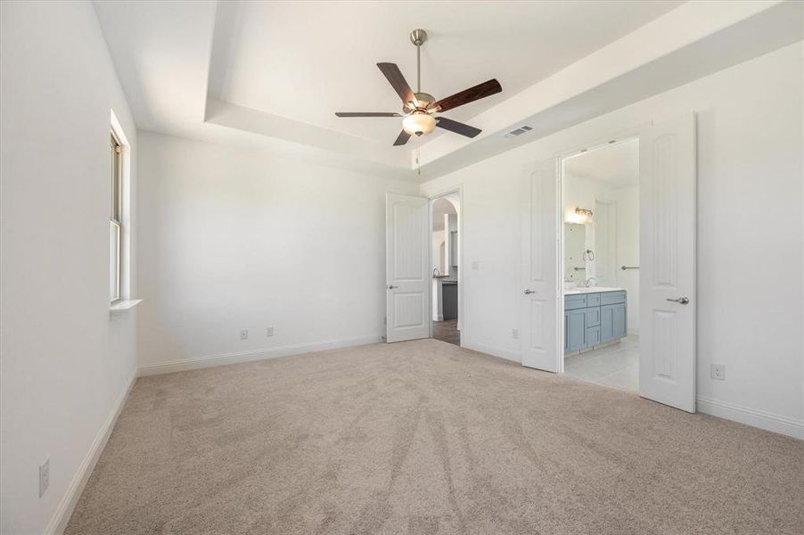 Unfurnished bedroom featuring ceiling fan, a raised ceiling, light carpet, and ensuite bath