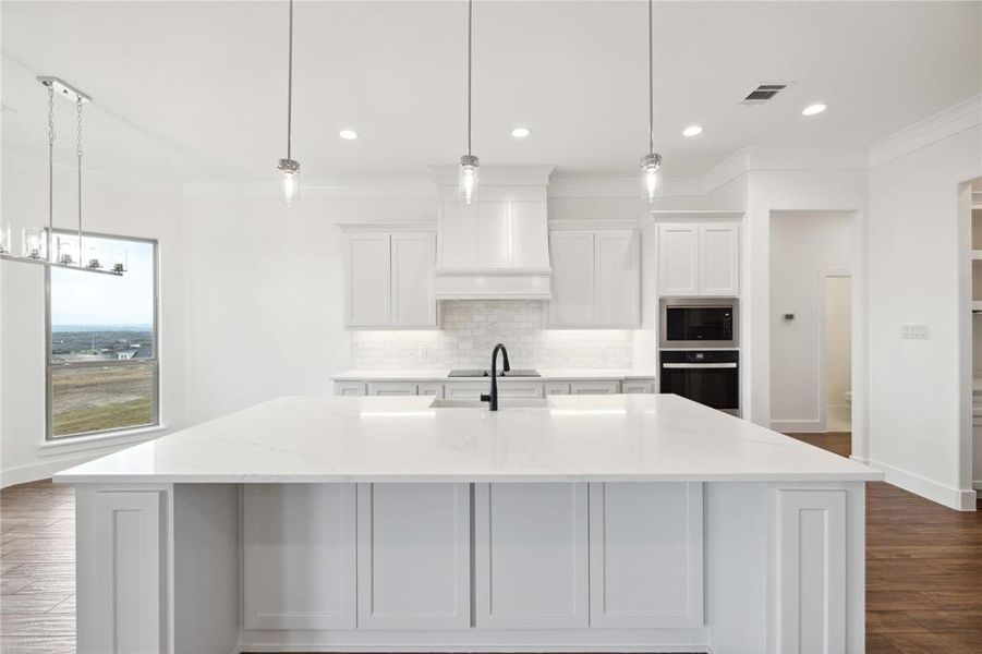 Kitchen with an island with sink, pendant lighting, custom exhaust hood, and light stone counters