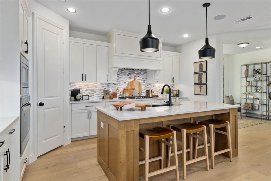 Kitchen featuring light wood-type flooring, hanging light fixtures, white cabinetry, light stone counters, and a kitchen island with sink