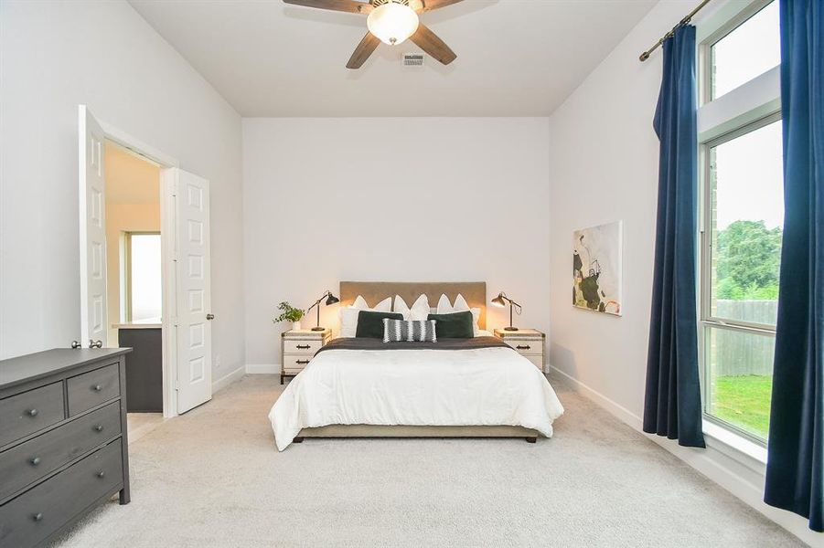 Well-lit primary bedroom featuring neutral walls, newly carpeted flooring, a ceiling fan, and large windows with a view of greenery outside. Also, includes a cozy sitting area for relaxation.