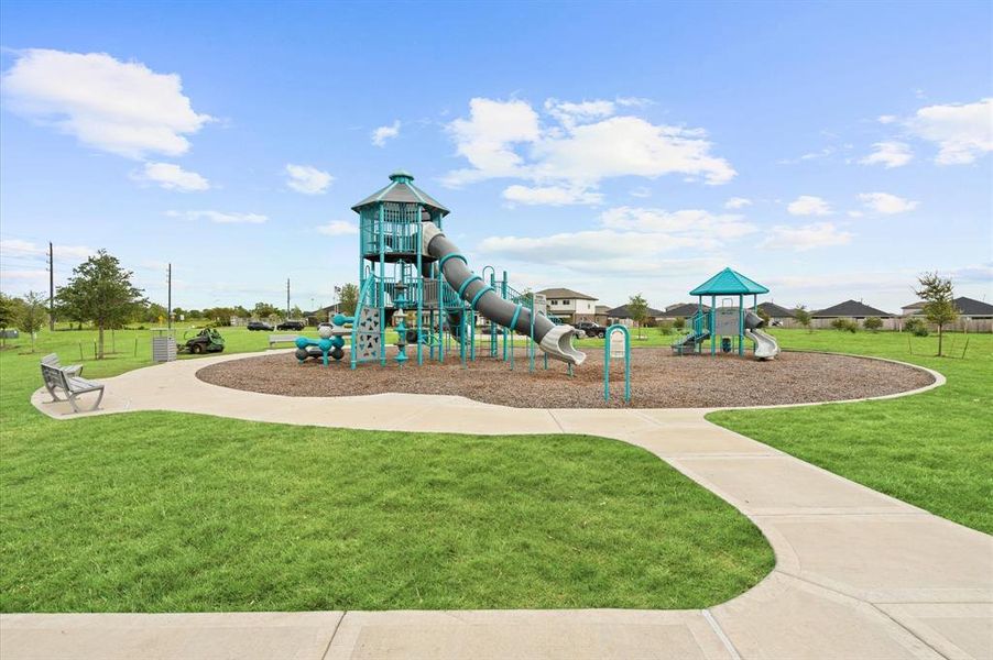 Community playground with modern equipment and safety surfacing, set within a well-maintained grassy area with a walking path and bench, offering a recreational space for families.