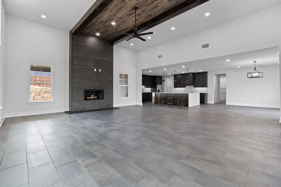 Unfurnished living room with a fireplace, a high ceiling, ceiling fan with notable chandelier, beam ceiling, and light tile patterned floors