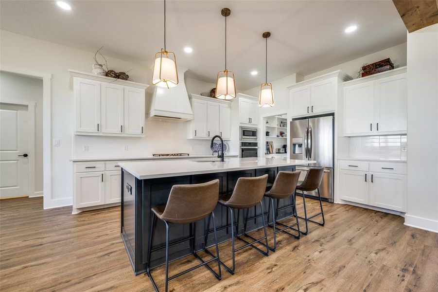 Kitchen with custom range hood, light hardwood / wood-style floors, backsplash, a kitchen island with sink, and appliances with stainless steel finishes