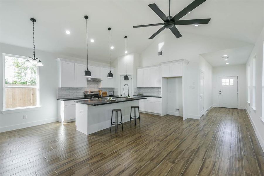 This is a spacious, modern kitchen with white cabinetry, stainless steel appliances, a large island with a sink, and elegant pendant lighting. The room features dark hardwood floors, high ceilings with a ceiling fan, and plenty of natural light from windows and a back door.