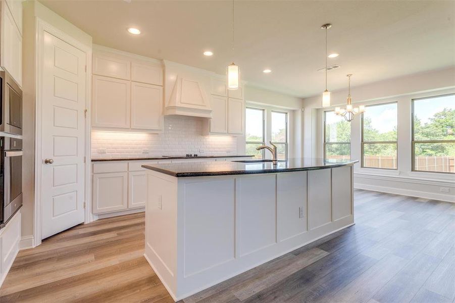 Kitchen with tasteful backsplash, a kitchen island with sink, custom exhaust hood, light wood-type flooring, and white cabinets