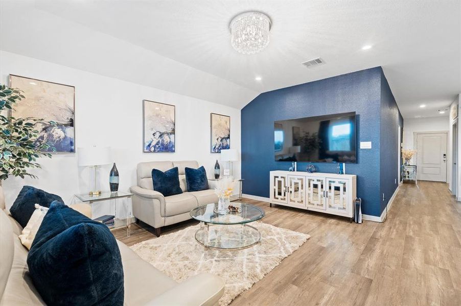 This is a modern and spacious living room with elegant premium laminate floors and a striking blue accent wall. It features a crystal chandelier, contemporary furnishings, and tasteful artwork, creating a stylish and comfortable space. Approximate Measurements: 16x14