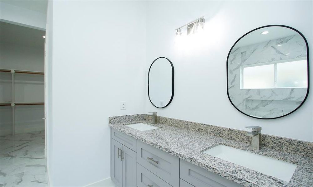 Modern bathroom with sleek finishes, featuring a double vanity with granite countertops, undermount sinks, and oval mirrors. There's a glimpse of a spacious walk-in closet, and the room is completed with marble-style tile flooring.