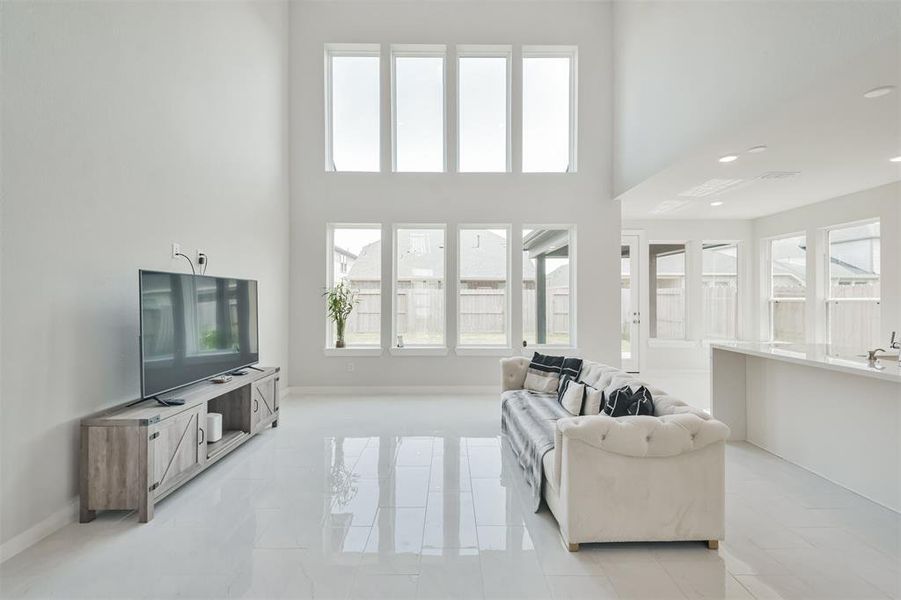 This image showcases a bright and airy living space with high ceilings and large windows allowing for ample natural light. The room features a modern aesthetic with sleek, glossy floor tiles and a neutral color palette.