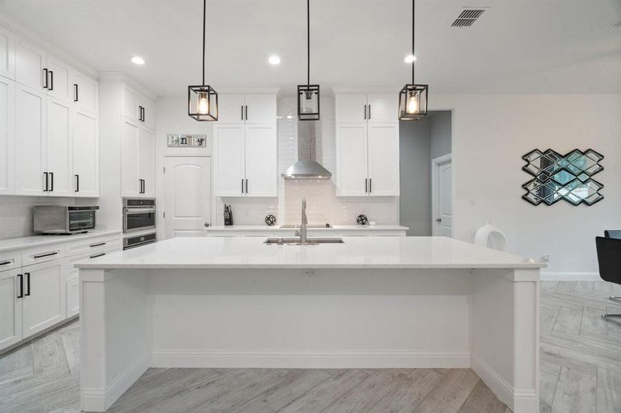 Kitchen with an island with sink, light wood-type flooring, hanging light fixtures, and tasteful backsplash