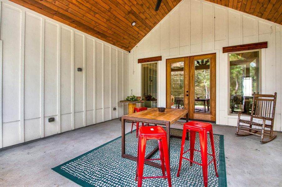 A cozy, rustic-style the covered back patio featuring a wooden ceiling, white walls with built-in storage, and a large glass door providing ample natural light and views of the surrounding greenery.