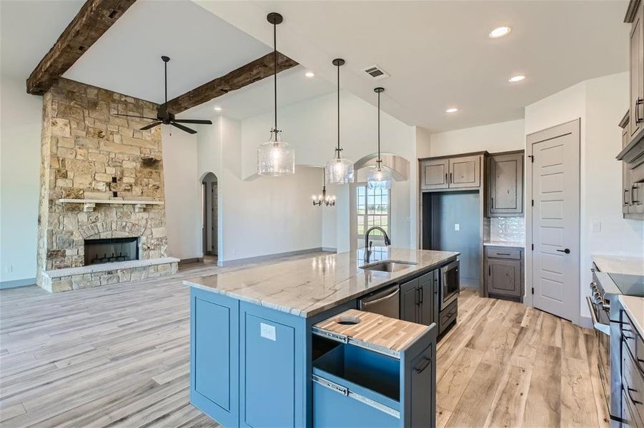 Kitchen featuring light hardwood / wood-style flooring, a center island with sink, a stone fireplace, beam ceiling, and ceiling fan