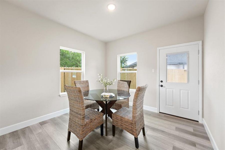 This is a bright, cozy dining space featuring a modern table with wicker chairs, sleek wood-look flooring, ample natural light from two windows, and a view of the backyard through a glass-paned door.