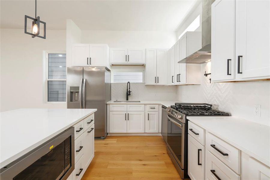 This is a modern kitchen featuring white cabinetry, stainless steel appliances, a gas stove, a herringbone tile backsplash, and light wood flooring. There's also an island with space for seating and a stylish pendant light above. Lots of natural light from 3 windows in the kitchen area.