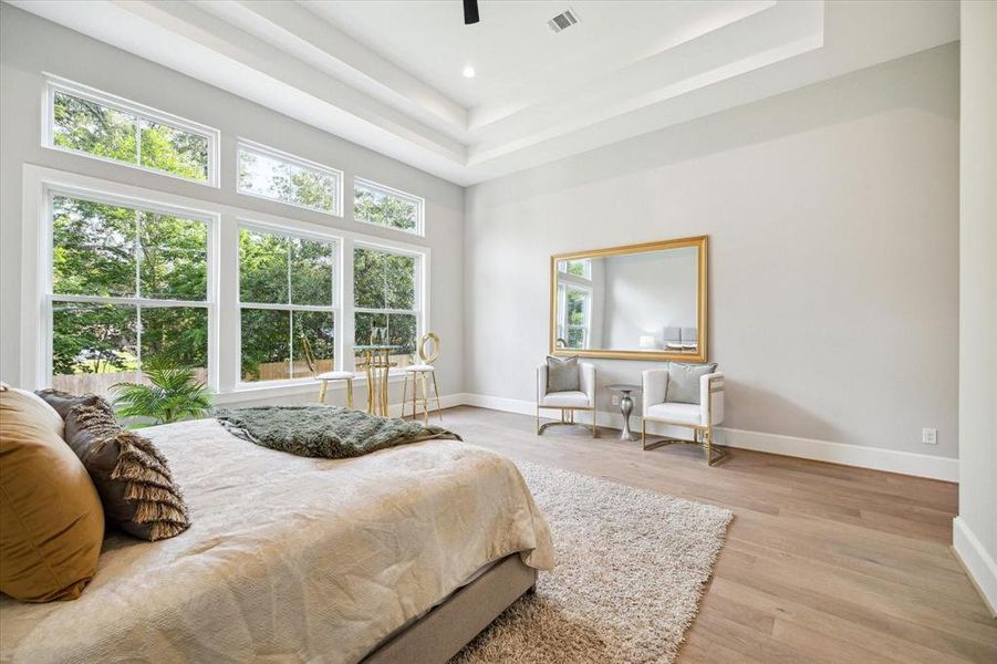 This is a spacious and well-lit bedroom with high ceilings and large windows that offer a view of greenery outside. It features a neutral color palette, a cozy bed, and an elegant seating area with a mirror, creating a serene and inviting atmosphere.