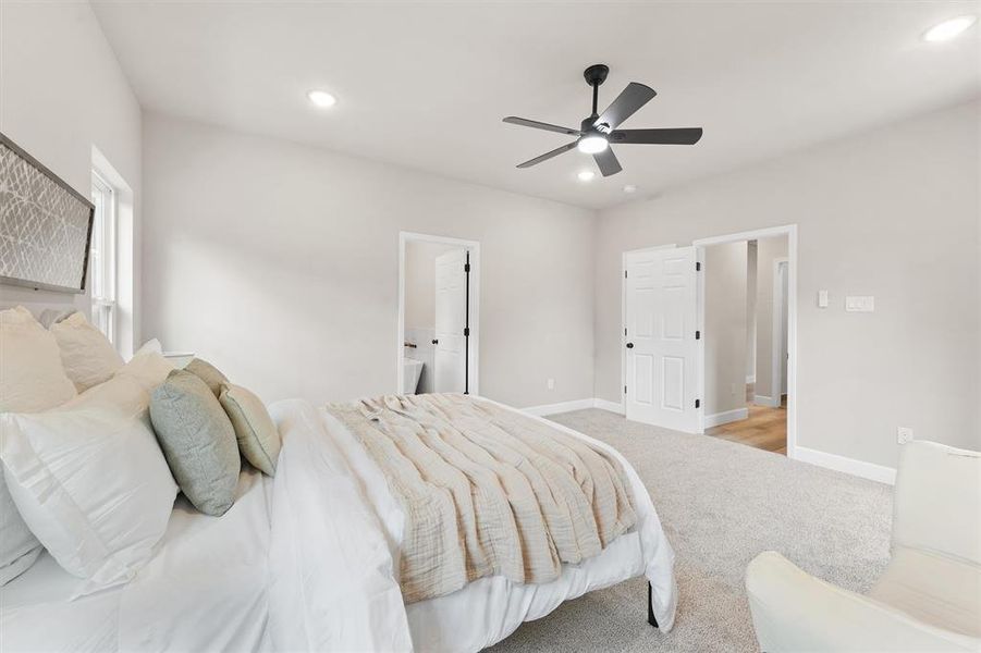 This is a spacious, well-lit bedroom featuring neutral colors, a modern ceiling fan with lights, plush carpeting, and two doors suggesting an en-suite bathroom and a closet or passage to another room.
