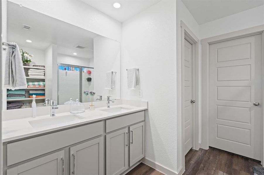 This is a modern, bright bathroom featuring dual sinks set in a spacious white vanity with ample storage. There's a large mirror above the vanity and an open shelving unit for towels and bathroom essentials. The room also includes a walk-in shower with a glass door and a separate toilet area behind a closed door. The flooring is a wood-look laminate, providing a nice contrast to the white walls and fixtures. Approximate Measurements:11X8