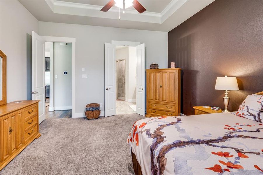 Bedroom with light colored plush carpet, en-suite bathroom, ceiling fan, and a tray ceiling