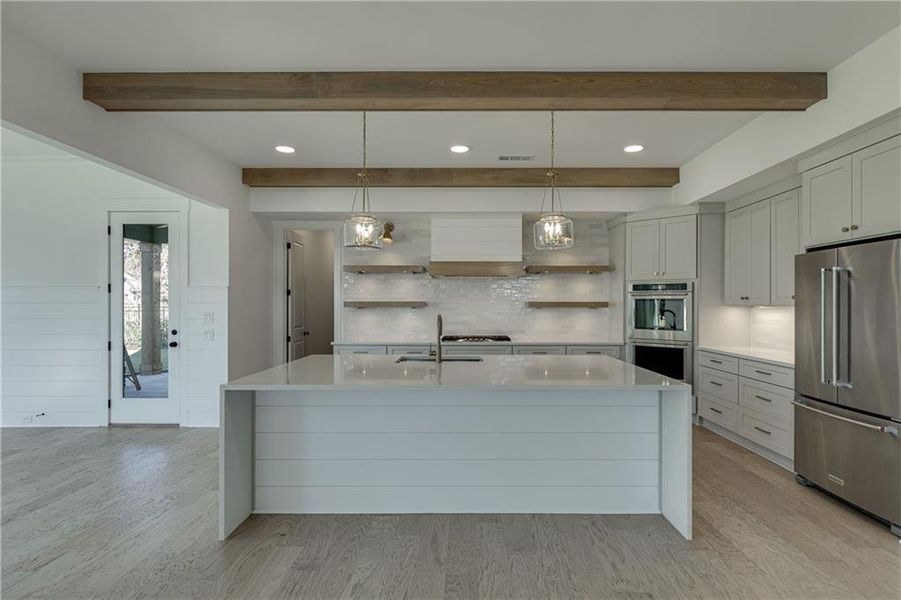 Kitchen with beamed ceiling, hanging light fixtures, appliances with stainless steel finishes, a kitchen island with sink, and light hardwood / wood-style flooring
