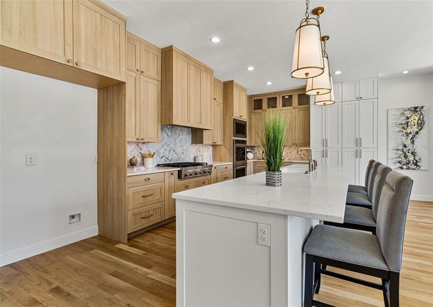 Kitchen featuring stainless steel appliances, a center island with sink, light stone countertops, pendant lighting, and light wood-type flooring