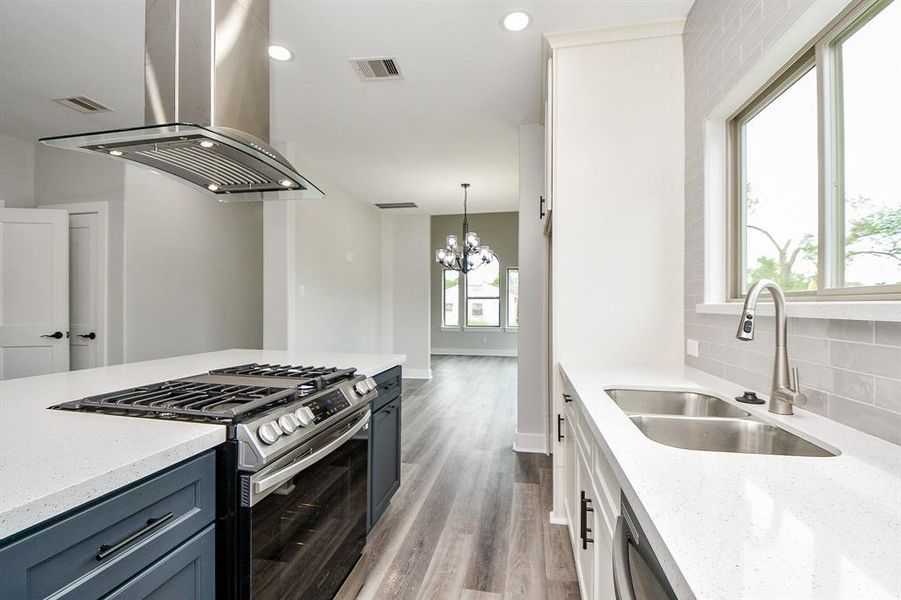 This is a modern, well-lit kitchen featuring stainless steel appliances, a gas range with a stylish overhead vent, dark cabinetry, light countertops, a subway tile backsplash, and a deep sink with a view from the window. The open layout leads to a spacious living area with abundant natural light.