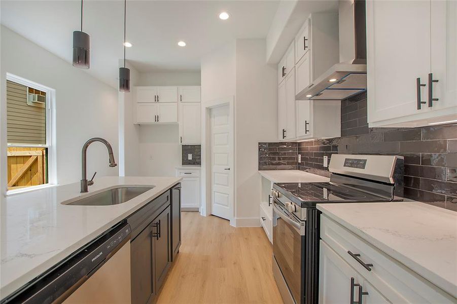 Kitchen with sink, light stone countertops, hanging light fixtures, appliances with stainless steel finishes, and wall chimney range hood