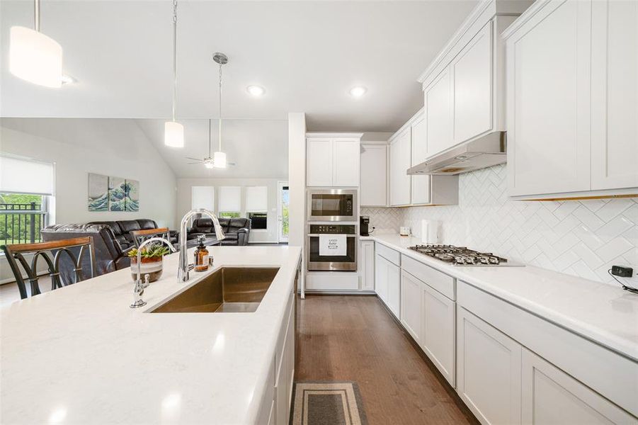 Open-plan kitchen featuring  a gas stove, and an island with a sink. The space is well-lit with natural and pendant lighting, and it flows into a dining area with a view outdoors.