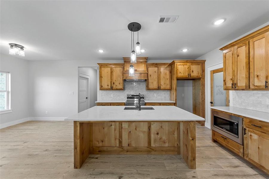 Kitchen with hanging light fixtures, appliances with stainless steel finishes, light hardwood / wood-style flooring, backsplash, and a kitchen island with sink