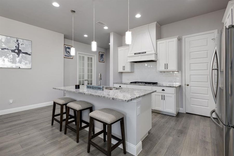 Kitchen featuring custom range hood, stainless steel fridge, a kitchen island with sink, and white cabinets