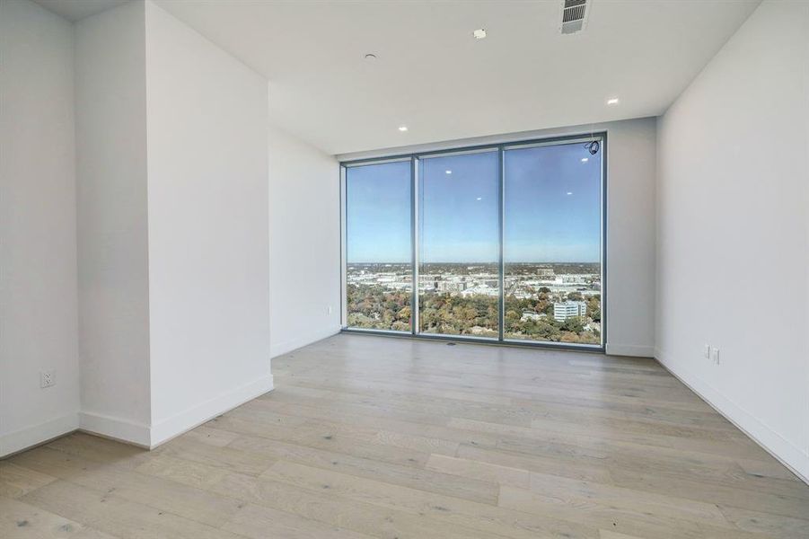 This is a bright, modern primary bedroom with large floor-to-ceiling windows offering expansive views of the surrounding cityscape. The room features light hardwood flooring and a clean, minimalistic design, ready for personalization.