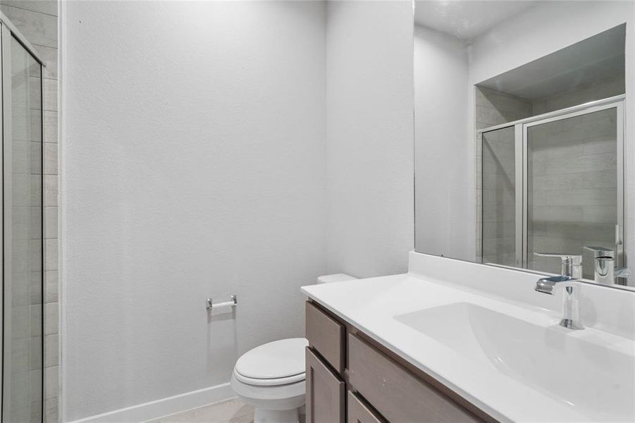 Secondary bath features tile flooring, shower with tile surround, dark stained cabinets, beautiful light countertops, mirror, dark, sleek fixtures and modern finishes.