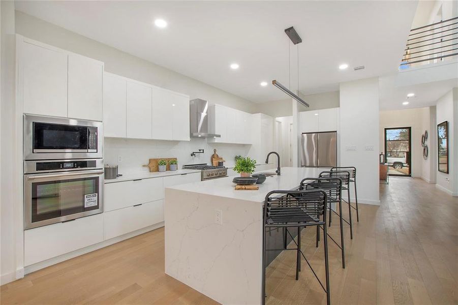 Kitchen with wall chimney range hood, stainless steel appliances, a center island with sink, pendant lighting, and white cabinets