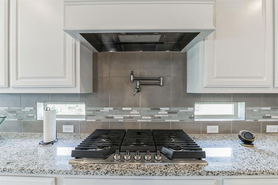 Kitchen featuring stainless steel gas cooktop, decorative backsplash, light stone countertops, white cabinetry, and custom range hood