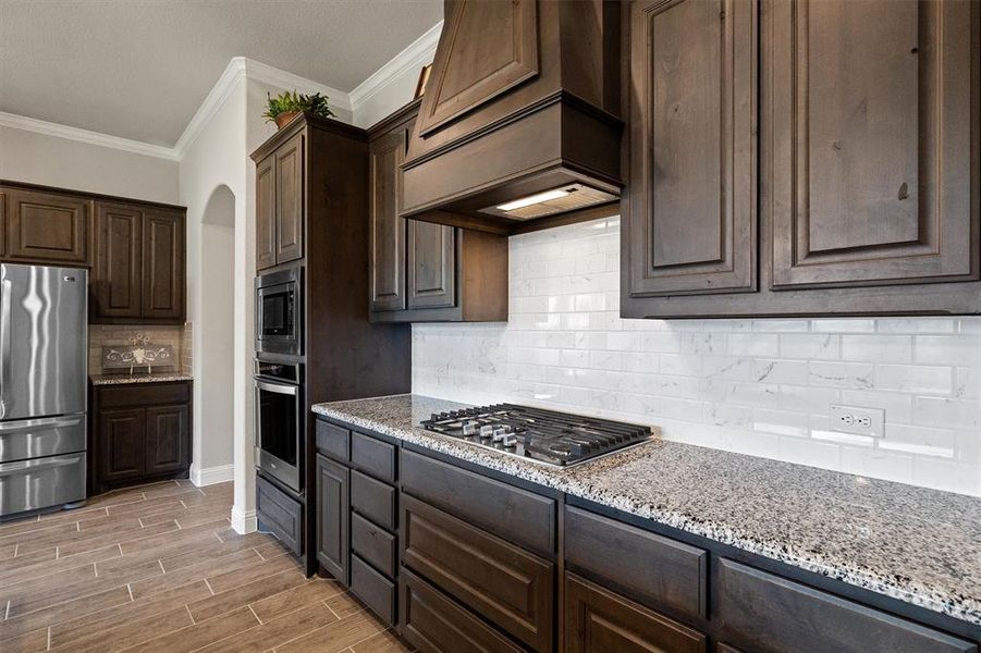 Kitchen featuring light stone counters, tasteful backsplash, crown molding, appliances with stainless steel finishes, and premium range hood