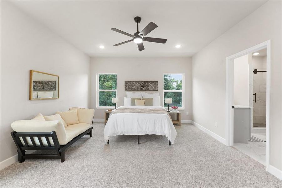 This is a spacious, well-lit bedroom featuring a large bed, a comfortable daybed, and an en-suite bathroom with a glass shower. The room has neutral colors, carpeted flooring, and is equipped with a ceiling fan. Two windows provide natural light and a view of greenery outside.