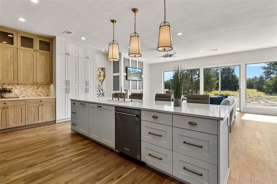 Kitchen featuring light brown cabinetry, wood-type flooring, dishwasher, pendant lighting, and a center island with sink