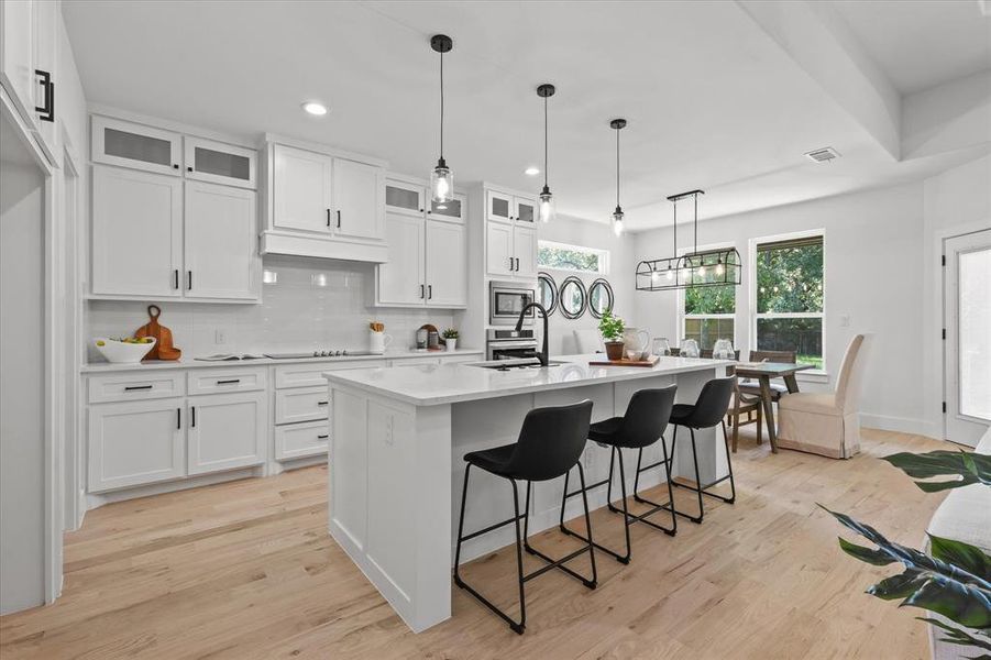 Kitchen with stainless steel microwave, a kitchen island with sink, hanging light fixtures, light wood-type flooring, and white cabinets