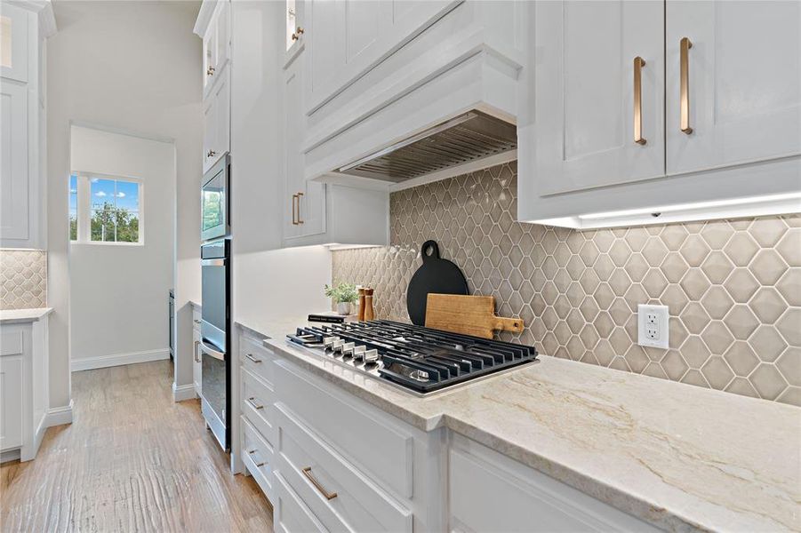 Kitchen with decorative backsplash, white cabinets, custom exhaust hood, appliances with stainless steel finishes, and light stone counters