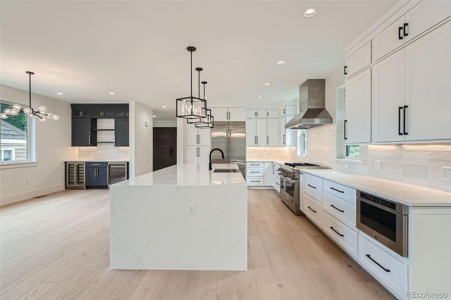 Chef's kitchen showcasing a massive quartz waterfall island, complemented by sleek quartz countertops, undercabinet lighting, stainless steel refrigerator/freezer, six-burner gas range, built-in hood vent, dishwasher, and microwave.