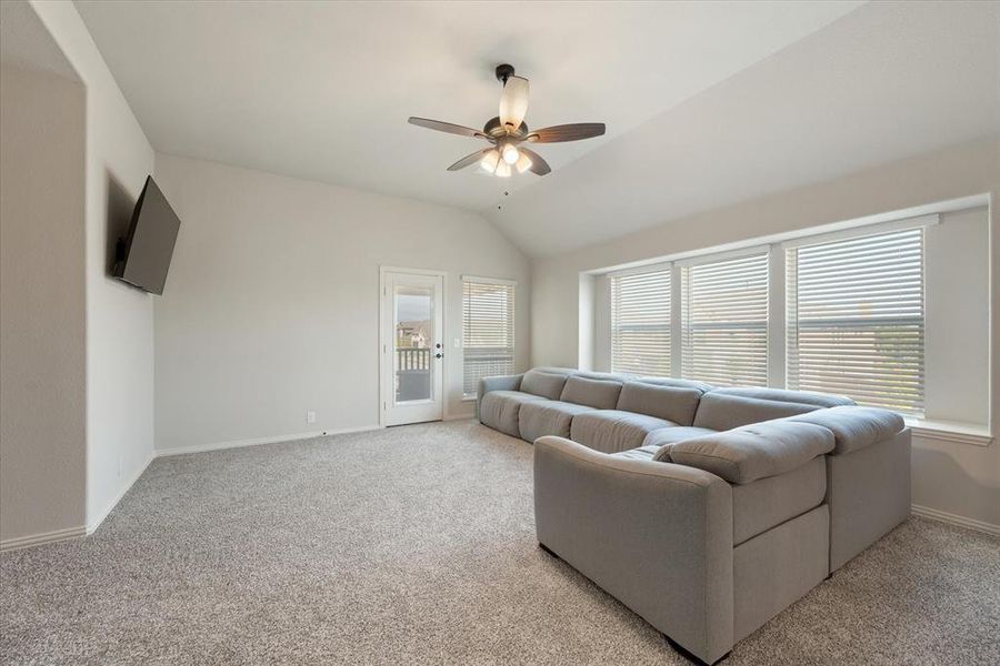 This upstairs game or second living room features a vaulted ceiling, ceiling fan with light kit, and light carpeting.  The door ahead leads to a covered balcony that overlooks the front yard.