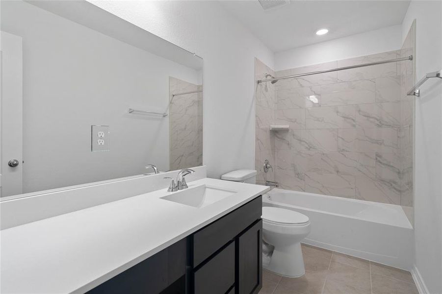 Secondary bath features tile flooring, bath/shower combo with tile surround, dark stained wood cabinets, beautiful light countertops, mirror, dark, sleek fixtures and modern finishes.