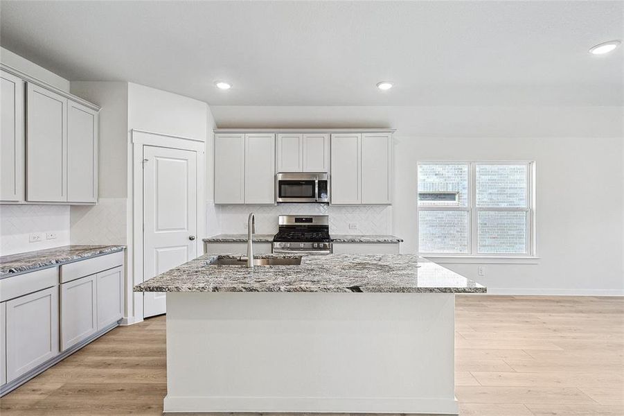 Kitchen featuring backsplash, light wood-type flooring, light stone counters, stainless steel appliances, and a kitchen island with sink