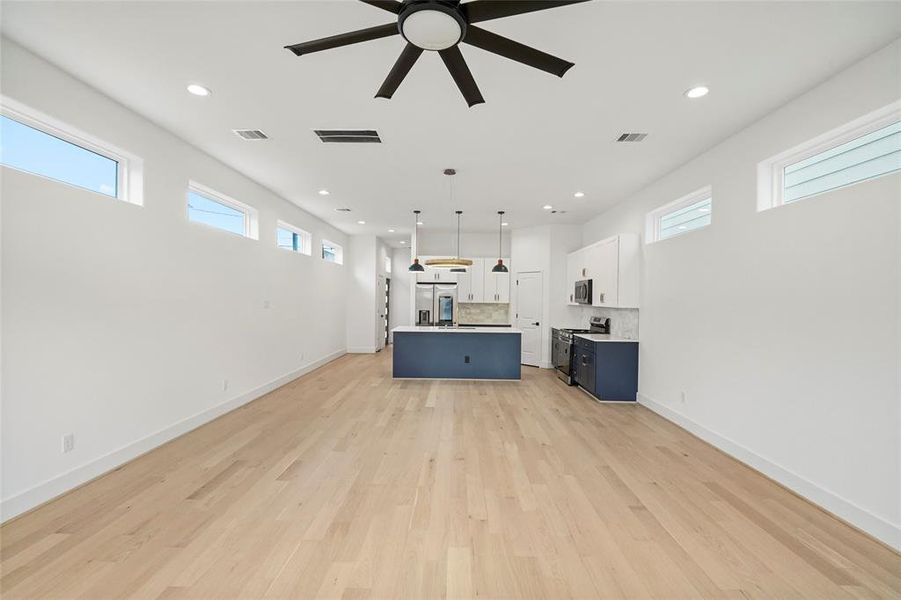 Modern open-plan living space with ample natural light, featuring a sleek kitchen with white cabinetry and a contrasting blue island. The room is finished with light hardwood floors and recessed lighting, creating an airy and contemporary feel.