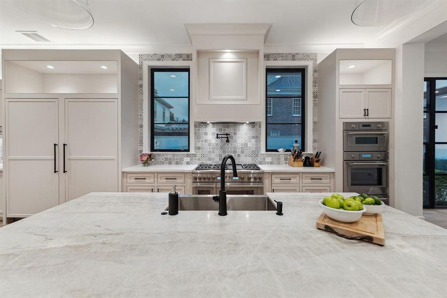 In this view, the stunning quartzite countertop is highlighted, accompanied by a stainless apron sink, a Riobel faucet, and a soap dispenser. This view shows the beautifully framed tall windows that flank the vent hood and provide a captivating view of the pool and spa.