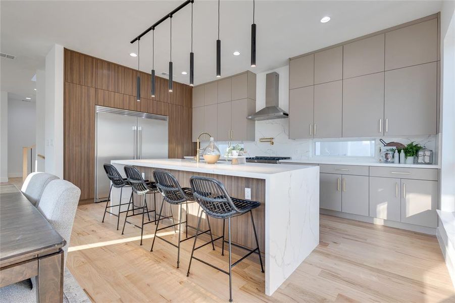 Kitchen with light wood-type flooring, decorative backsplash, wall chimney exhaust hood, hanging light fixtures, and a center island with sink