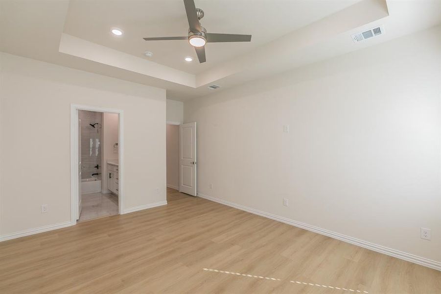 Unfurnished bedroom featuring a raised ceiling, ensuite bathroom, light hardwood / wood-style flooring, and ceiling fan