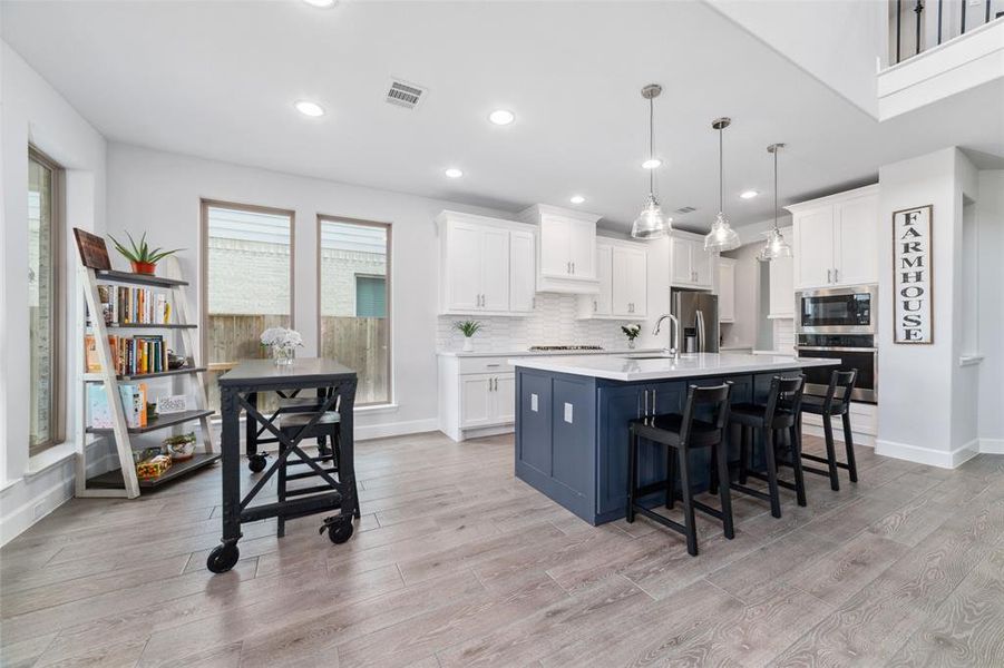 This is a spacious, modern kitchen featuring white cabinetry, a contrasting blue island with seating, stainless steel appliances, and elegant pendant lights. The room has a bright and airy feel with natural light from large windows and high ceilings.