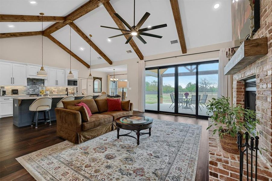 Living room with ceiling fan with notable chandelier, dark hardwood / wood-style flooring, a brick fireplace, beam ceiling, and high vaulted ceiling